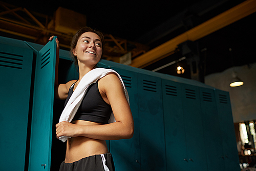 Dark low angle portrait of smiling young woman standing by locker in sports club and looking away, copy space