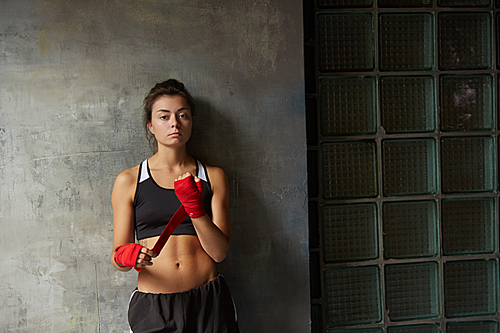 Waist up portrait of tough female fighter wearing hand wraps  while posing against concrete wall, copy space