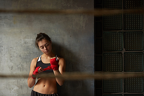 Waist up portrait of tough female fighter wrapping hands while standing against concrete wall in boxing club , copy space