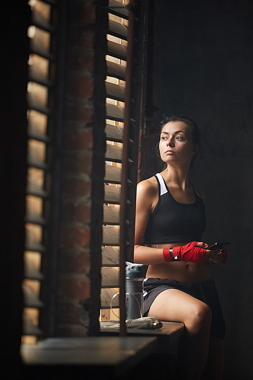 Portrait of young sportswoman sitting by window in dark room and looking away through shutters, copy space