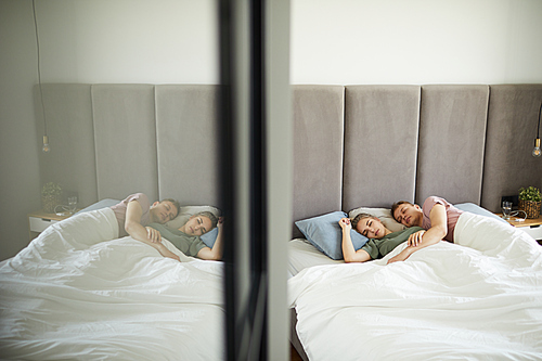 Young affectionate sleeping couple lying in bed under blanket by mirror on door of wardrobe