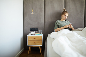 Young relaxed woman sitting in bed under banket while scrolling in smartphone