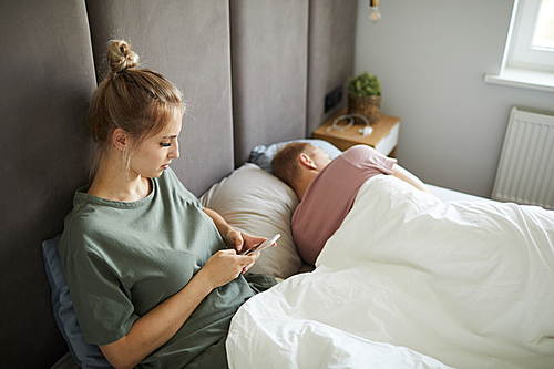 Suspicious young wife scrolling in her husband smartphone while sitting in bed