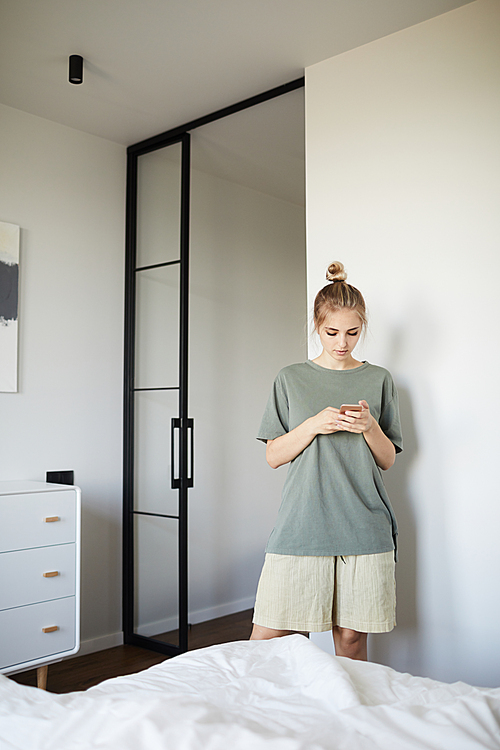 Pretty young casual woman scrolling in smartphone while standing by bed in bedroom