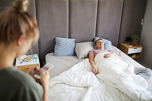 Young tired man sleeping in bed while wife or girlfriend scrolling in his smartphone near by