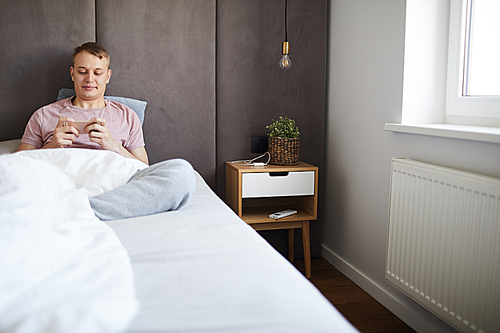 Happy relaxed guy sitting in bed under blanket and watching curious video in smartphone at leisure