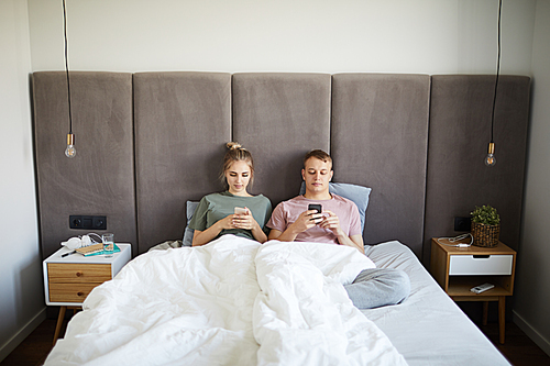 Young mobile couple lying in bed while using their smartphones after sleep