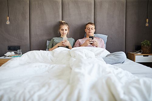 Young couple scrolling or playing in smartphones while spending leisure in bed