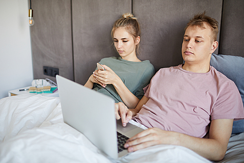 Young man with laptop networking in bed while his wife scrolling in smartphone