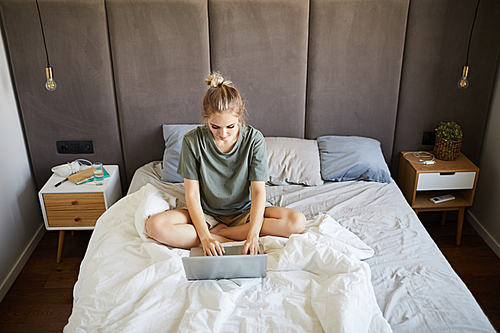 Pretty girl in casual t-shirt sitting on bed while typing on laptop keypad during network