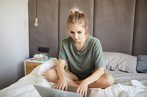 Cute blonde girl in t-shirt typing and looking at laptop display while sitting on bed in the morning