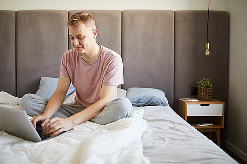 Young relaxed businessman in t-shirt networking in front of laptop in bed on weekend