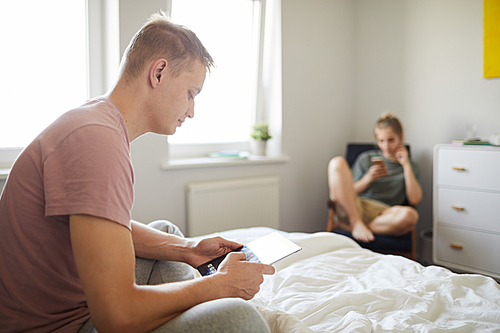 Contemporary guy with mobile gadget sitting on bed while scrolling in tablet with his wife on background