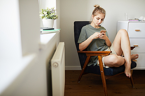 Cute girl in casualwear watching video in smartphone while relaxing in armchair in bedroom