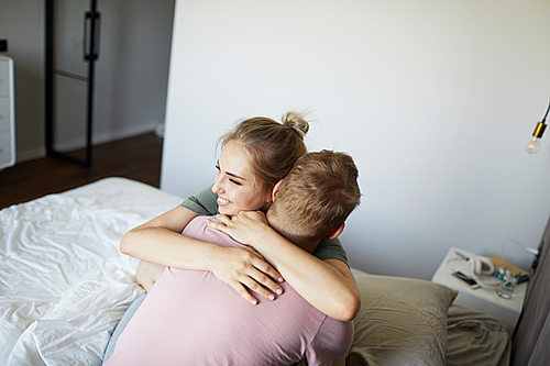 Cheerful young woman embracing her husband while both sitting on bed in the morning