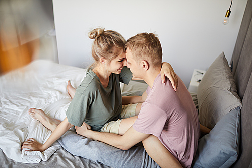 Happy young spouses in t-shirts sitting on bed in front of one another in embrace