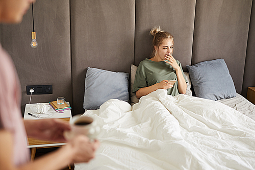 Young yawning woman with smartphone sitting in bed in the morning while her husband bringing her coffee