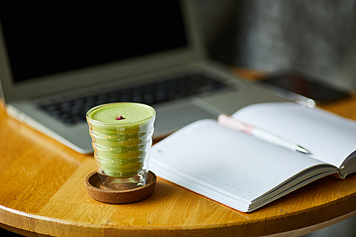 Small glass cup of bright green matcha tea on table with opened textbook and laptop