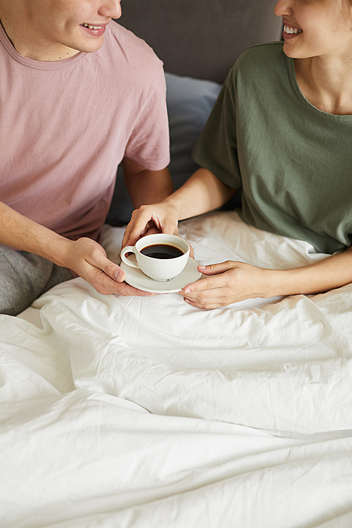 Happy young wife taking cup of coffee from husband while sitting under blanket in bed