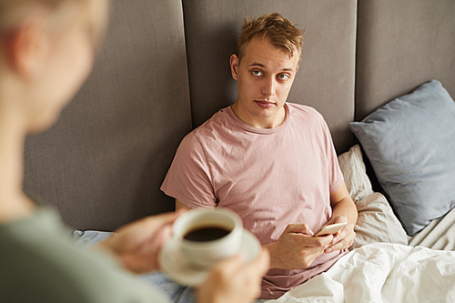 Sick or tired young man with smartphone looking at his wife bringing him cup of coffee in bed