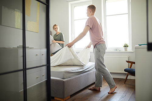 Young man in sweatpants and t-shirt doing bed while helping his wife in the morning after sleep