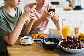 Served table with various fresh snacks and drinks and young couple using smartphones by breakfast