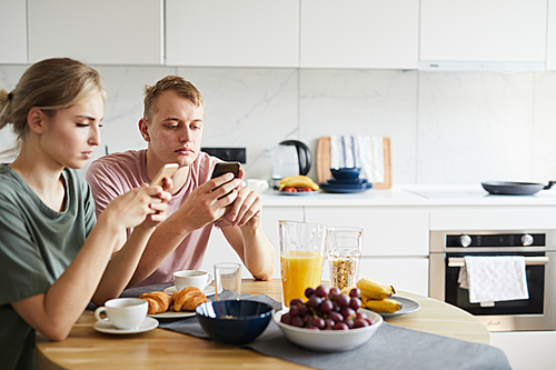 Young contemporary couple searching in social networks while sitting by served table in the kitchen
