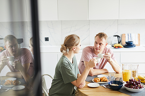 Young couple having tea or coffee by served table and talking during breakfast in the kitchen