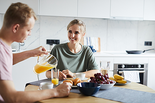 Young man pouring fresh orange juice in glass of his wife during breakfast in the kitchen