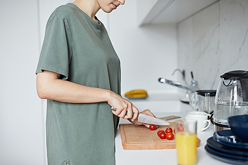 Young housewife with knife cutting red tomatoes on wooden board while preparing vegetable salad