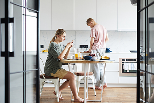 Young wife texting in smartphone by brunch while her husband cooking in the kitchen