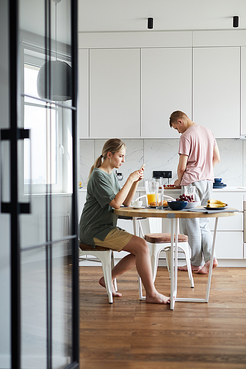 Young woman sitting by table in the kitchen, having breakfast and using smartphone while her husband cooking
