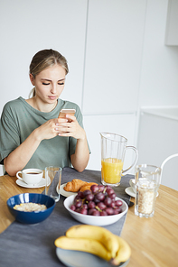 Young restful female watching video in smartphone by served table in the morning