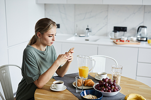 Young attractive woman using smartphone while having breakfast or brunch in the kitchen