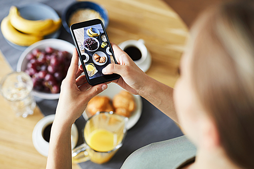 Contemporary girl with smartphone taking shot of her served table and the food prepared for breakfast