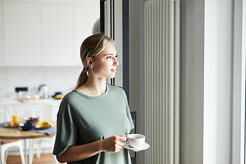 Young casual woman with cup of coffee looking through window while staying at home on weekend
