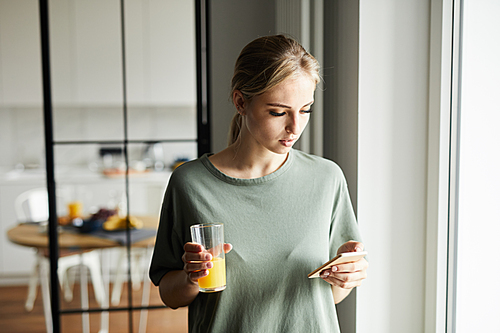 Pretty girl with glass of orange juice watching curious staff in smartphone while staying at home