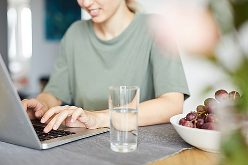 Young woman sitting by kitchen table at home and typing on laptop keypad during network