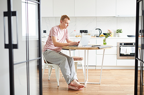 Young casual man sitting by table in the kitchen in front of laptop while networking
