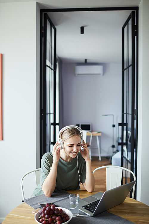 Happy young female in headphones sitting by kitchen table at home and interacting through video-chat