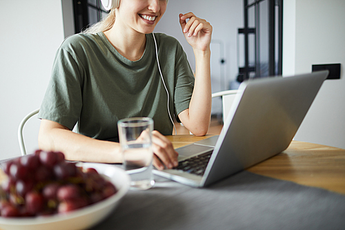 Smiling young woman sitting by table in front of laptop, networking or interacting in video-chat