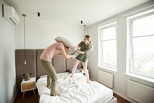 Young playful couple beating each other with pillows while standing on bed in the morning