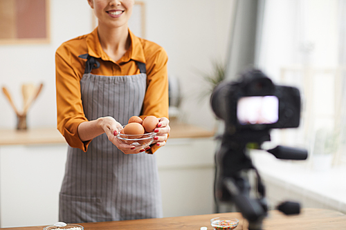 Cropped portrait of beautiful young woman holding eggs to camera while filming baking tutorial in studio, copy space