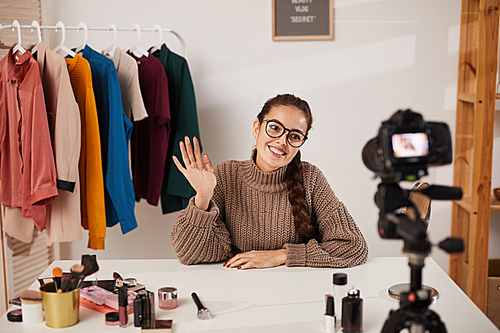 Portrait of smiling young woman waving at camera while filming video for beauty and lifestyle channel, copy space
