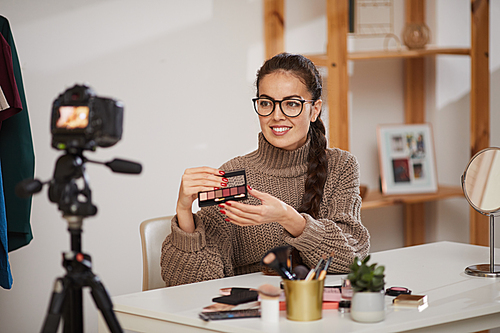 Portrait of smiling young woman showing make up products to camera while filming video review for beauty and lifestyle channel, copy space