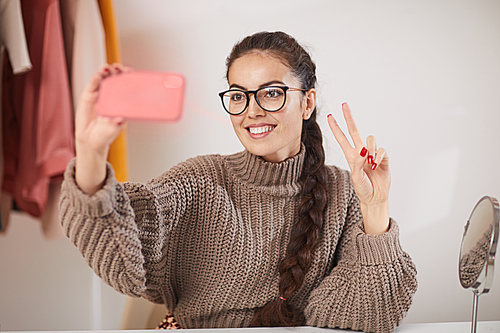 Portrait of contemporary young woman wearing glasses taking selfie photo via smartphone while sitting against plain white background in studio, copy space