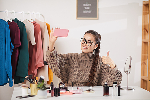 Portrait of contemporary young woman wearing glasses taking selfie photo via smartphone and showing thumbs up in beauty and makeup studio, copy space