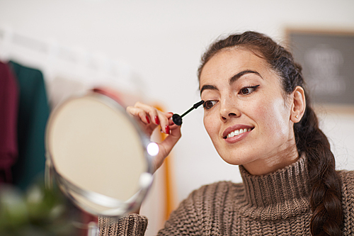 Portrait of smiling young woman applying mascara to eyelashes while doing make up at home, copy space
