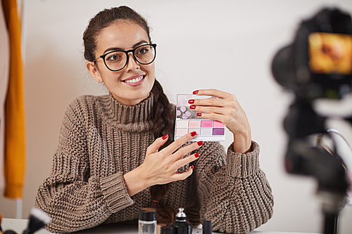 Portrait of contemporary young woman holding eyeshadow palette and smiling at camera while filming video review or tutorial for beauty and lifestyle channel, copy space