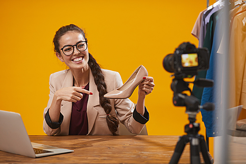 Portrait of contemporary young woman holding elegant shoe while filming video review for fashion and beauty channel against yellow background, copy space
